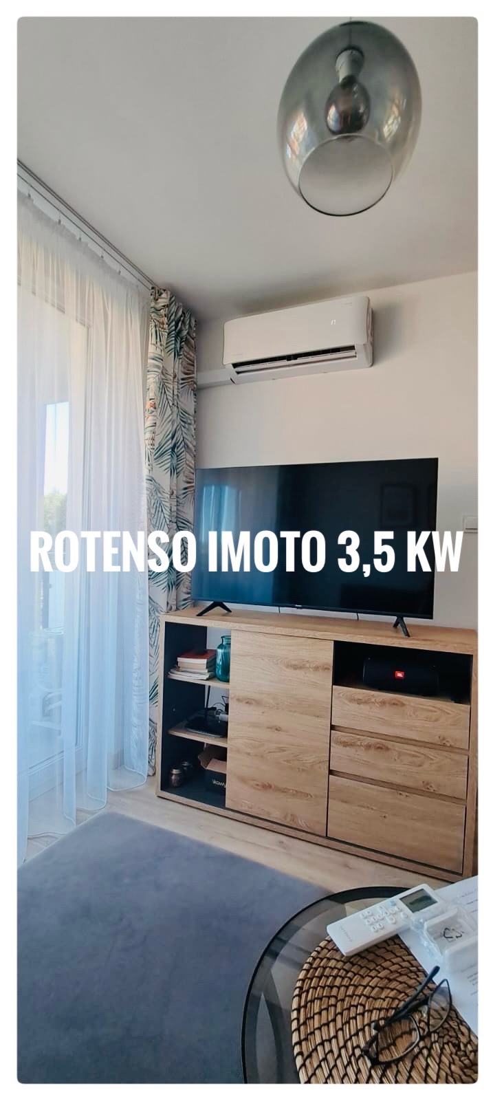 Biała jednostka klimatyzacyjna zamontowana nad drewnianą szafką RTV w salonie z zasłonami w tropikalne liście i szklanym stolikiem kawowym