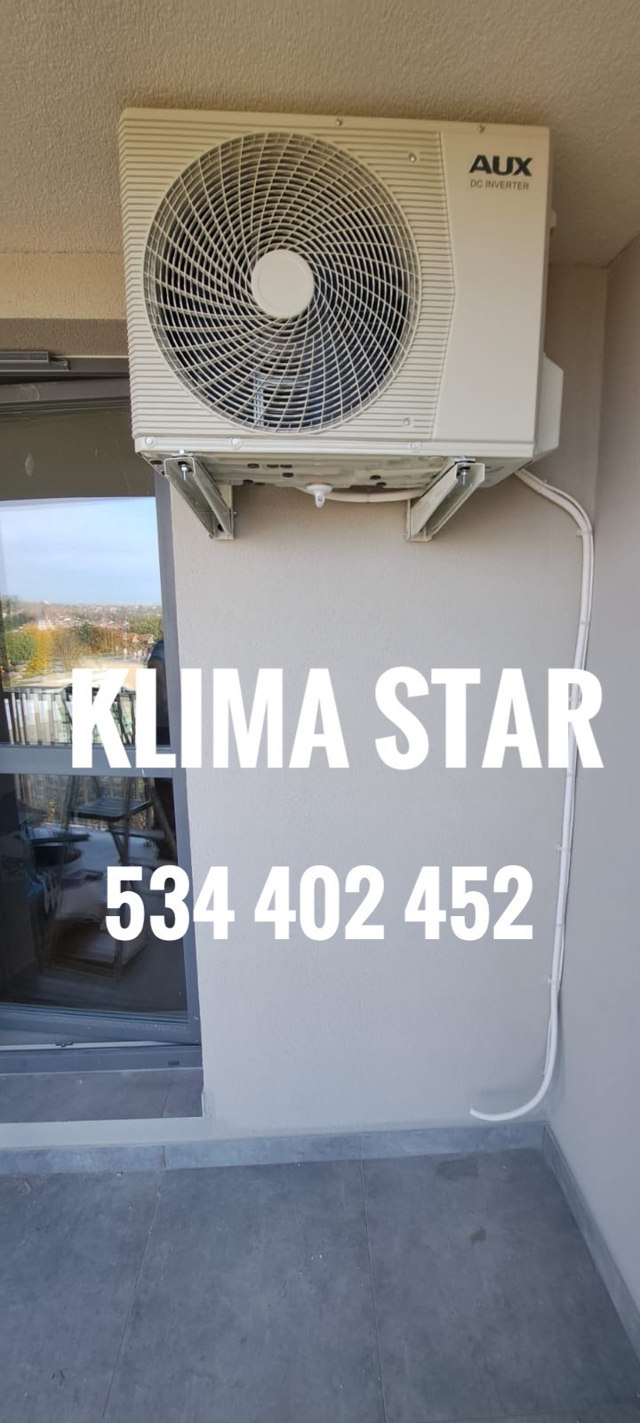 Zewnętrzna jednostka klimatyzacji AUX DC Inverter zamontowana na ścianie balkonu, widoczne białe kable prowadzące w dół ściany.