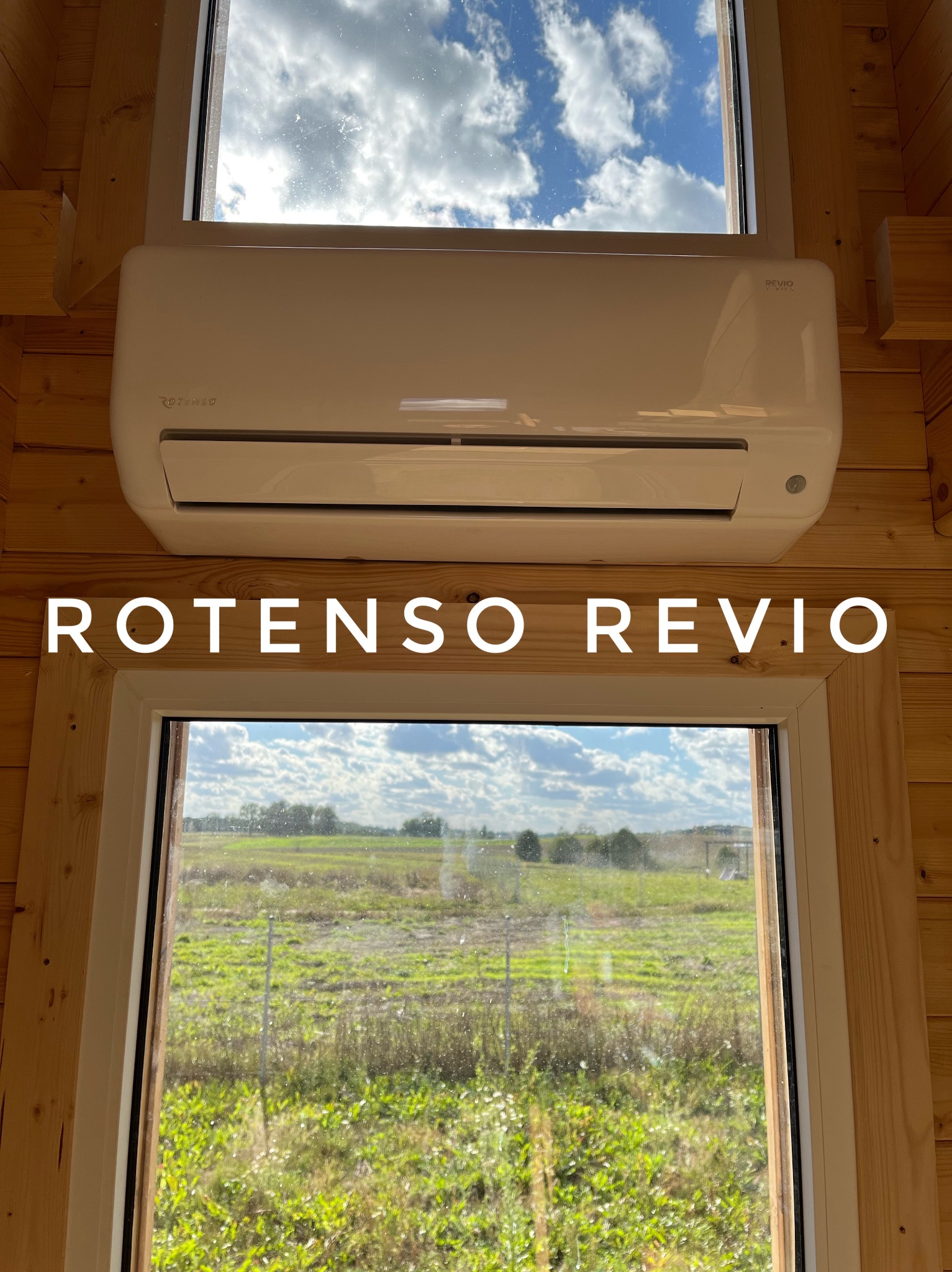 Biały klimatyzator Rotenso Revio zamontowany nad oknem w drewnianym domku, widok na zielone pole i zachmurzone niebo.