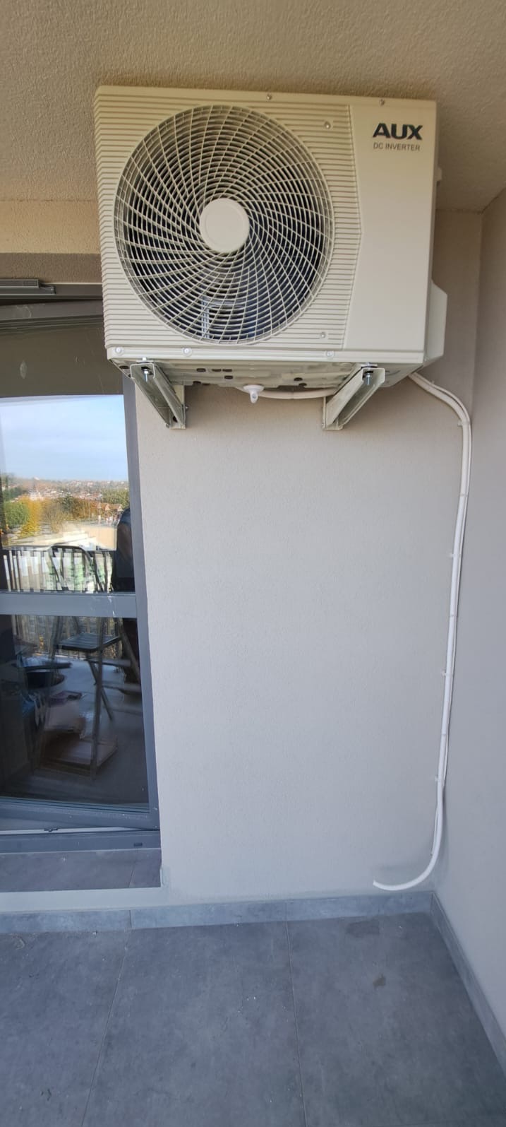 Zewnętrzna jednostka klimatyzacji AUX DC Inverter zamontowana na balkonie, widoczne białe kable prowadzące wzdłuż ściany.