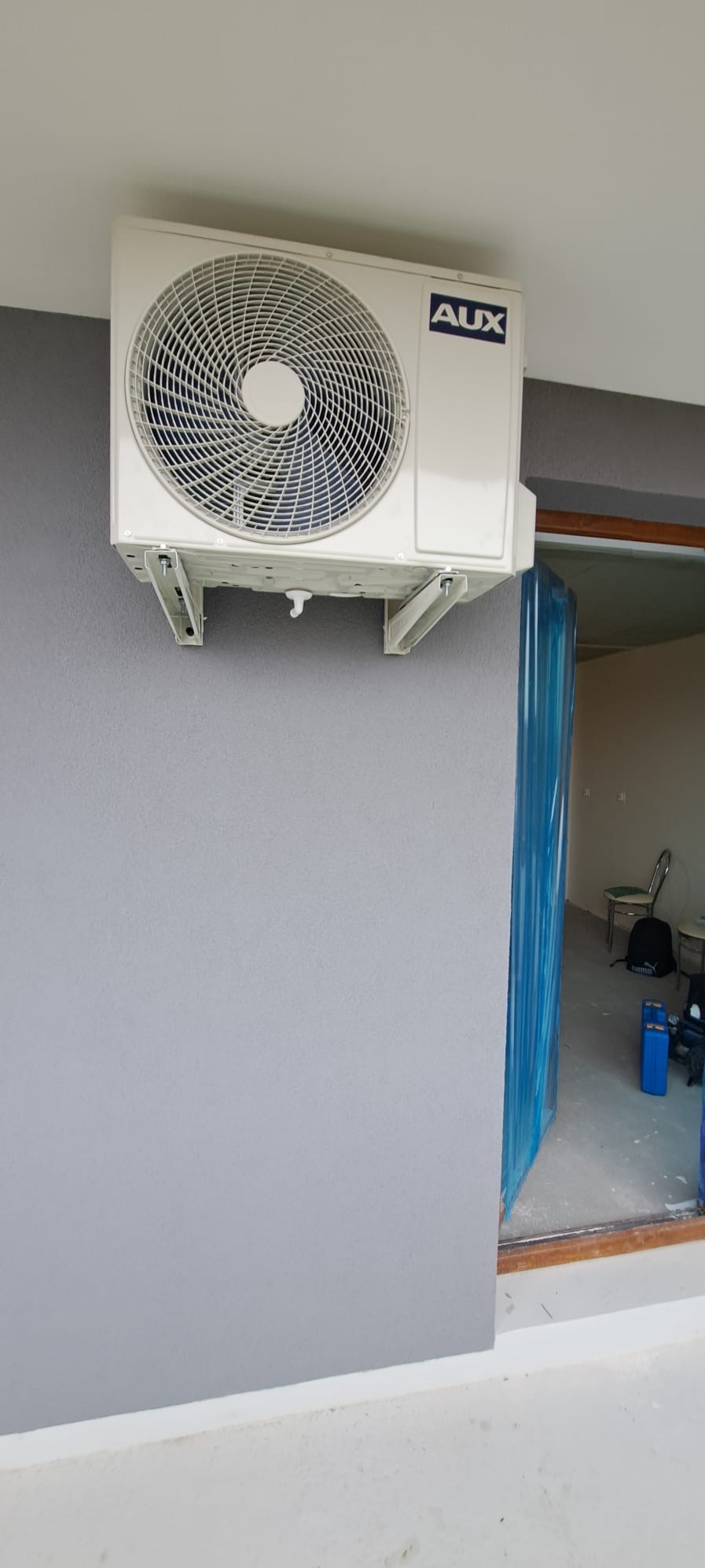 Biały klimatyzator marki AUX zamontowany na szarej ścianie wewnątrz budynku, widoczne metalowe wsporniki i fragment okna zabezpieczonego folią ochronną.