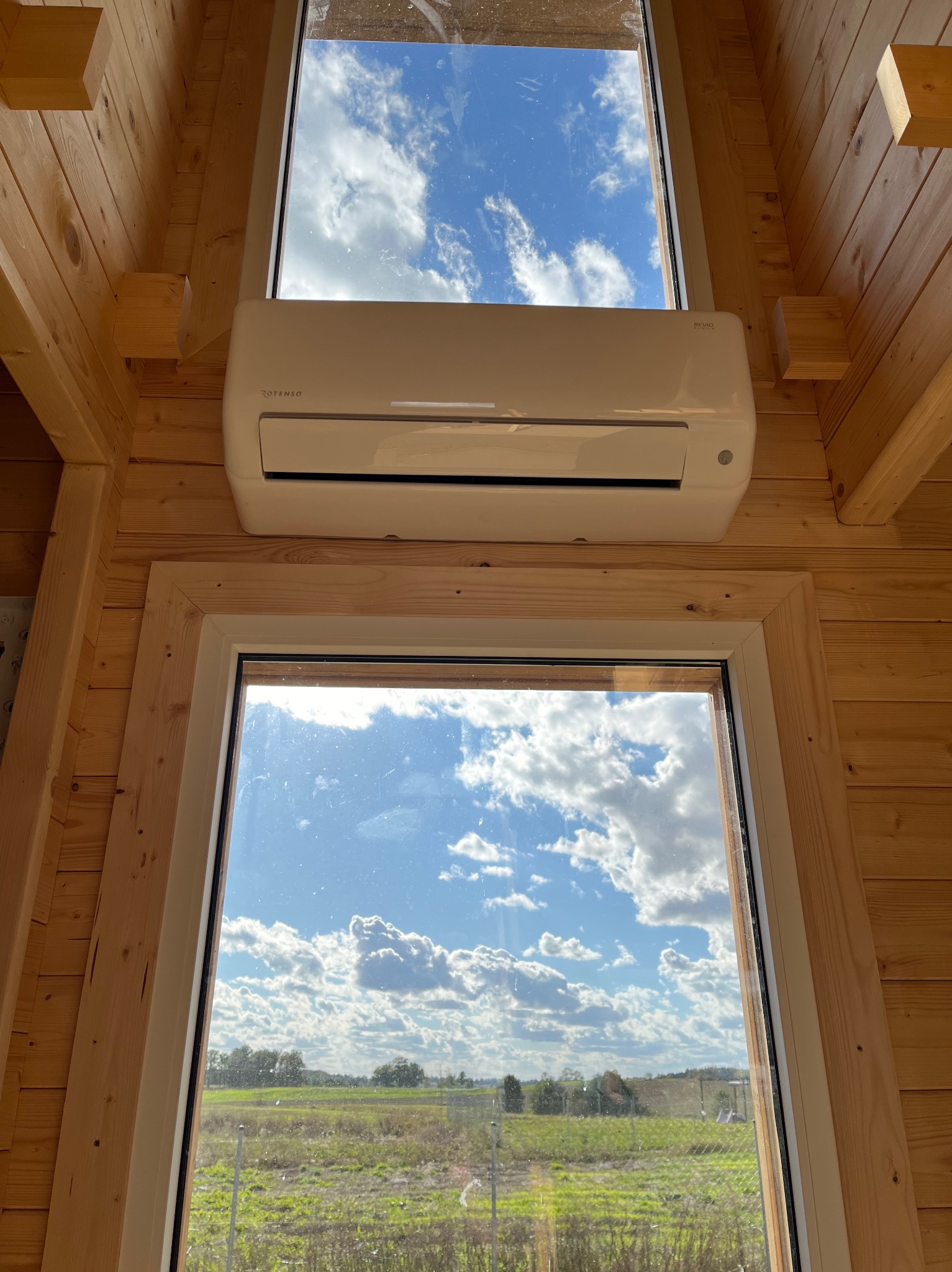 Wnętrze drewnianego domu z zamontowanym klimatyzatorem pod oknem, widok na pole i błękitne niebo z chmurami przez okna.