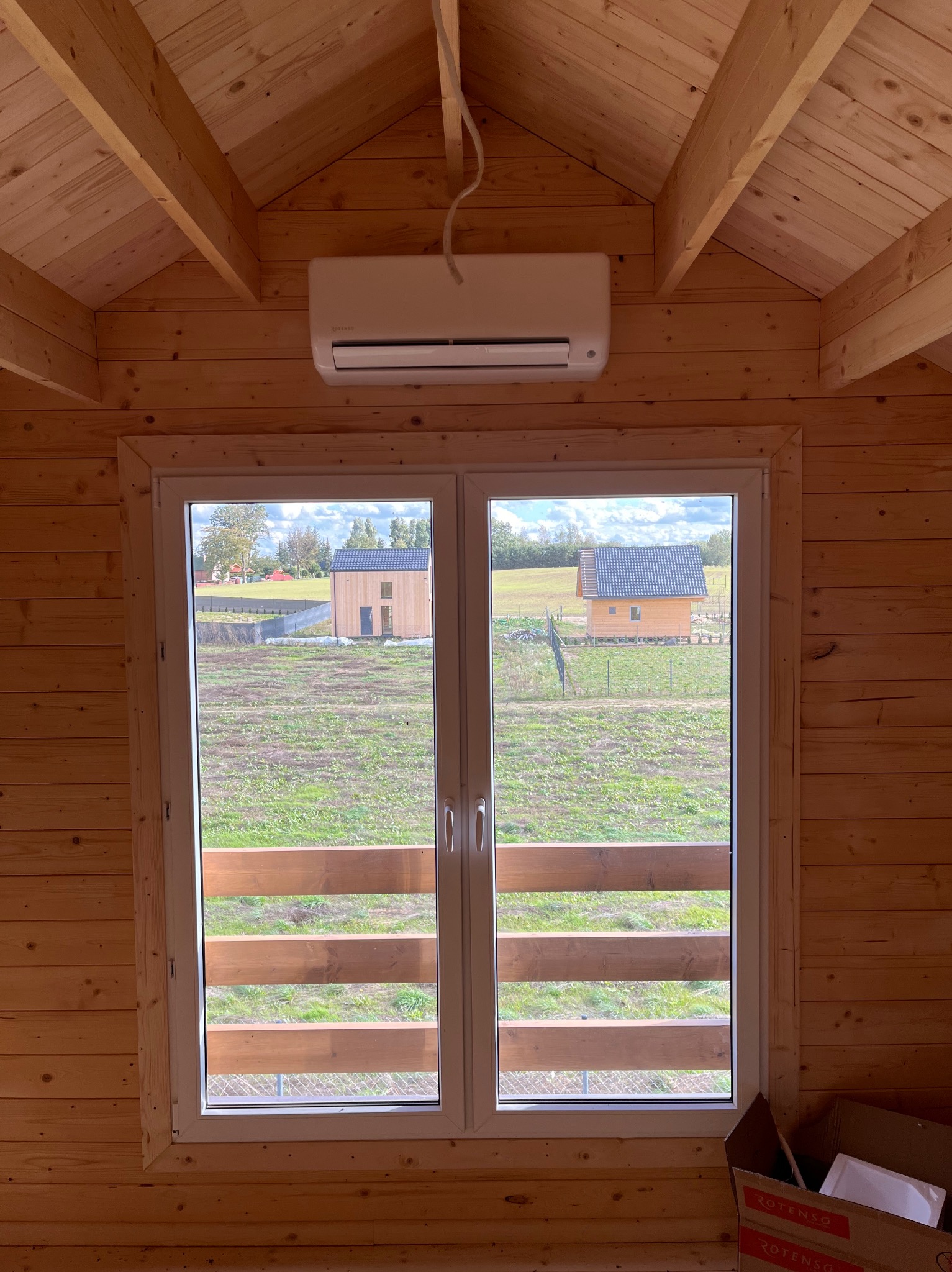 Wnętrze drewnianego domku z zamontowaną klimatyzacją ścienną nad oknem z widokiem na dwa inne domki.