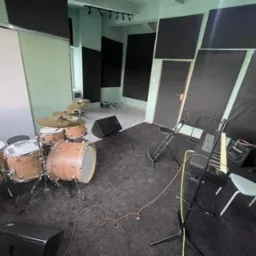 Wnętrze studia nagrań z zestawem perkusyjnym o jasnym wykończeniu drewna, pianinem elektrycznym i panelami akustycznymi na ścianach.