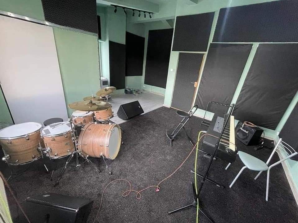 Wnętrze studia nagrań z zestawem perkusyjnym o jasnym wykończeniu drewna, pianinem elektrycznym i panelami akustycznymi na ścianach.