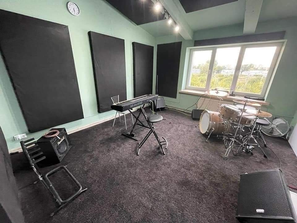 Wnętrze pokoju z pianinem M-Audio, perkusją, panelami akustycznymi na ścianach i oknem z widokiem na zieleń.