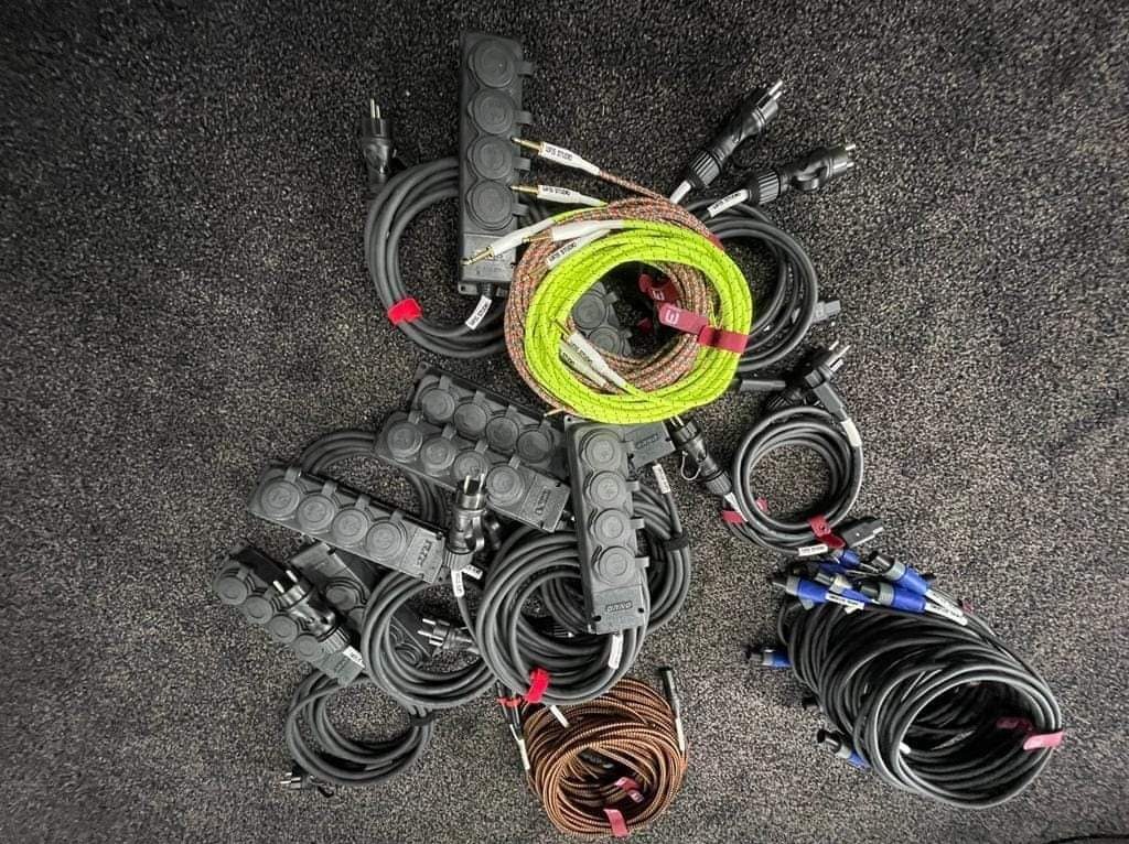 Zestaw kabli audio z wtyczkami i rozgałęźnikami na szorstkiej powierzchni, zwinięte w pętle i zabezpieczone taśmą, kable w kolorach czarnym, zielonym, brązowym i z niebieskimi wtyczkami.