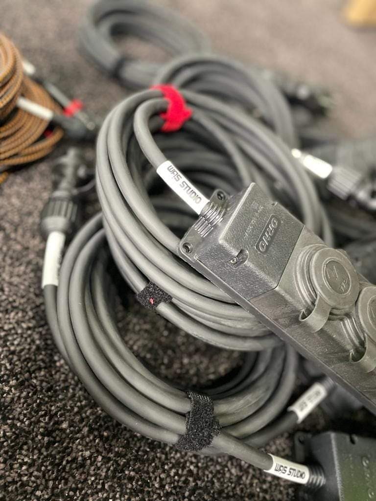 Zwinięte szare kable audio z czarnymi złączami i rzepami mocującymi, etykieta 'WAS STUDIO' na kablu, widoczne inne kable w tle.