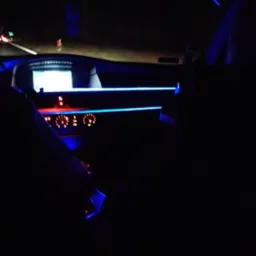 Wnętrze samochodu z podświetleniem LED w kolorze niebieskim, widoczne deska rozdzielcza, zegary i fragment przedniej szyby w ciemności.