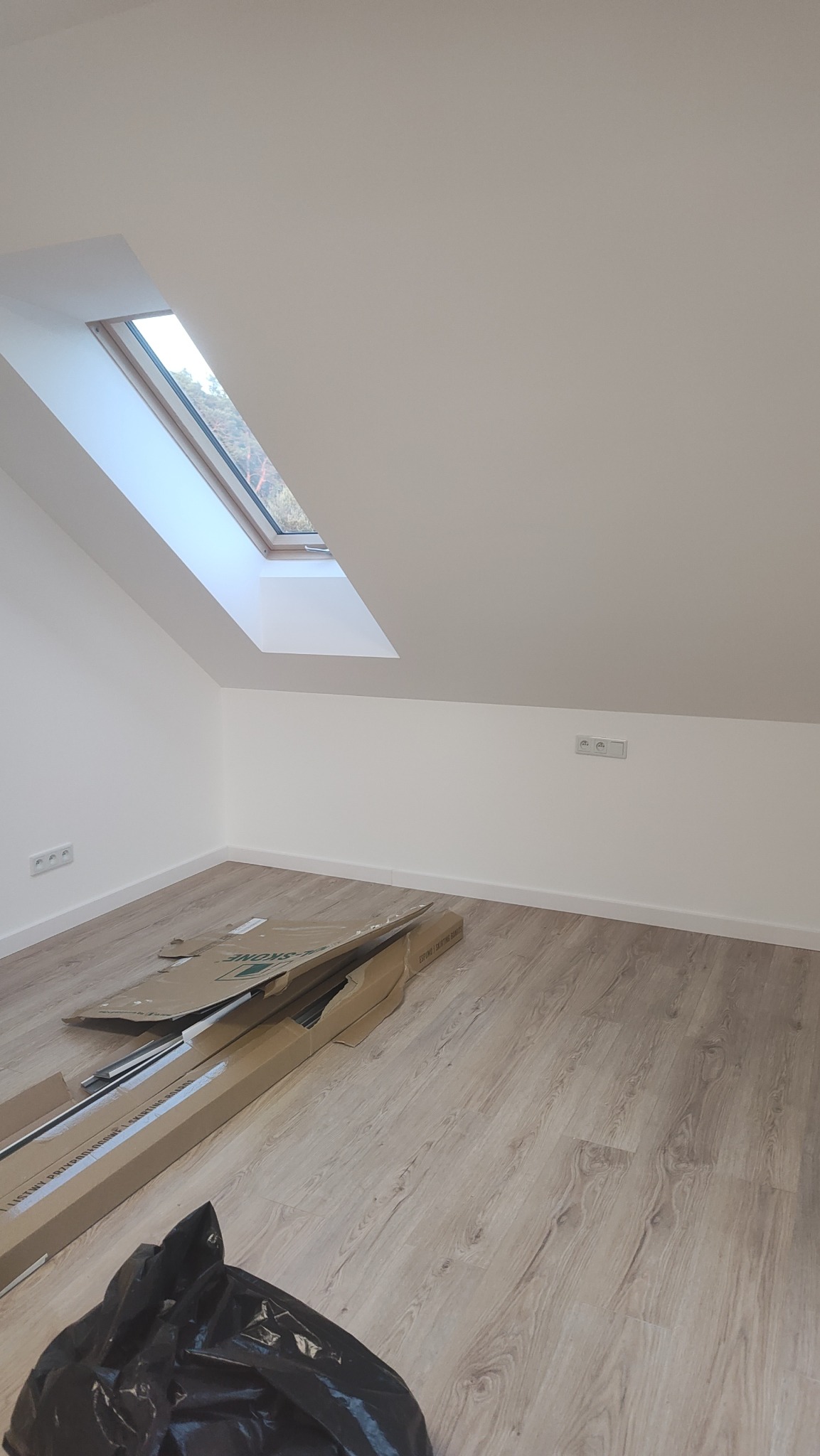 Pokój na poddaszu z oknem dachowym i panelami podłogowymi. Widoczne kartony po roletach okiennych i worki na śmieci. Świeżo pomalowane ściany na biało.
