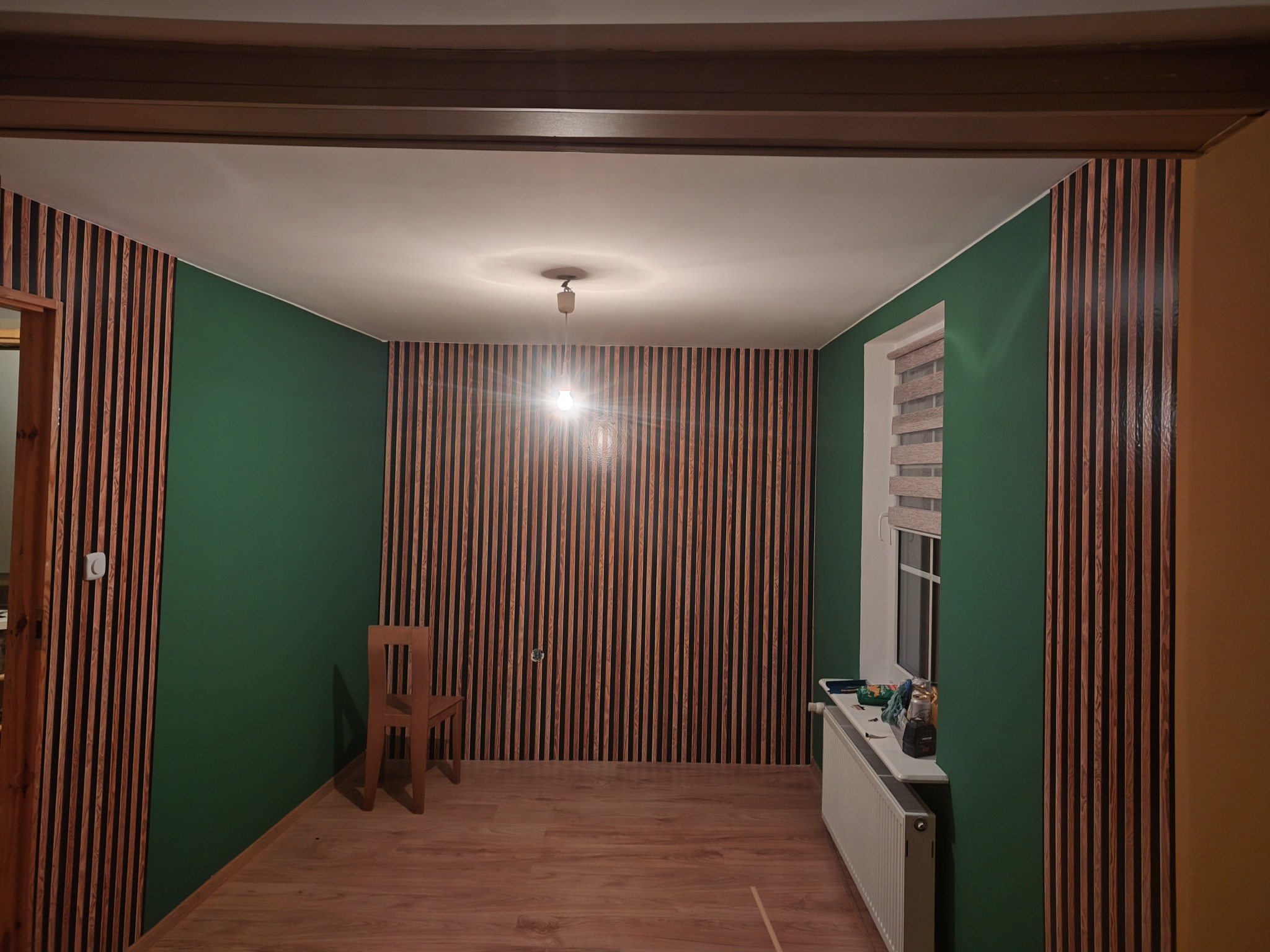 Pokój po remoncie: ściany w kolorze butelkowej zieleni i panele drewniane na ścianie z oknem. Na podłodze panele podłogowe imitujące drewno. Krzesło drewniane.