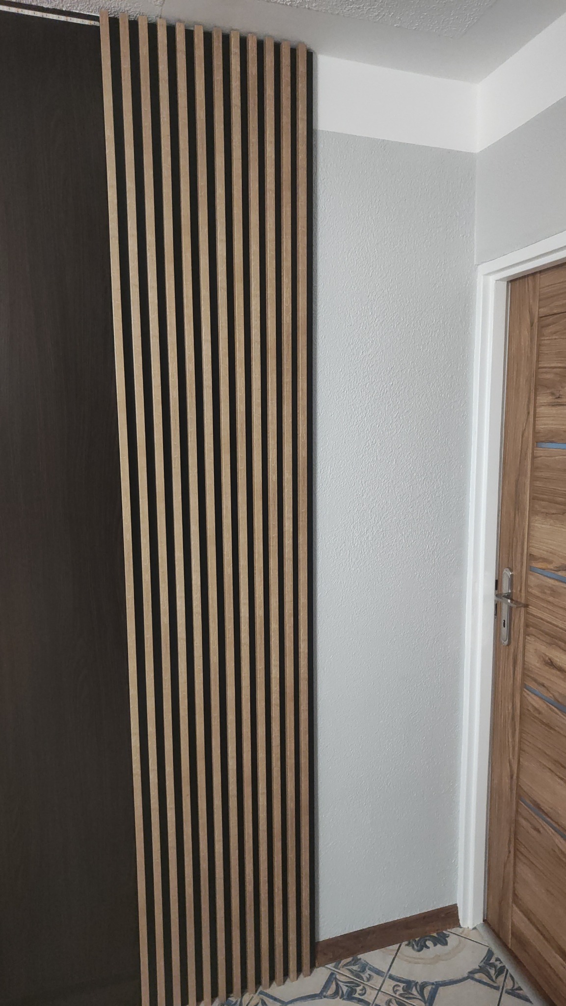 Panel lamelowy z drewnianymi listwami na ścianie, obok ciemnej szafy i drewnianych drzwi. Widoczna podłoga z wzorem.