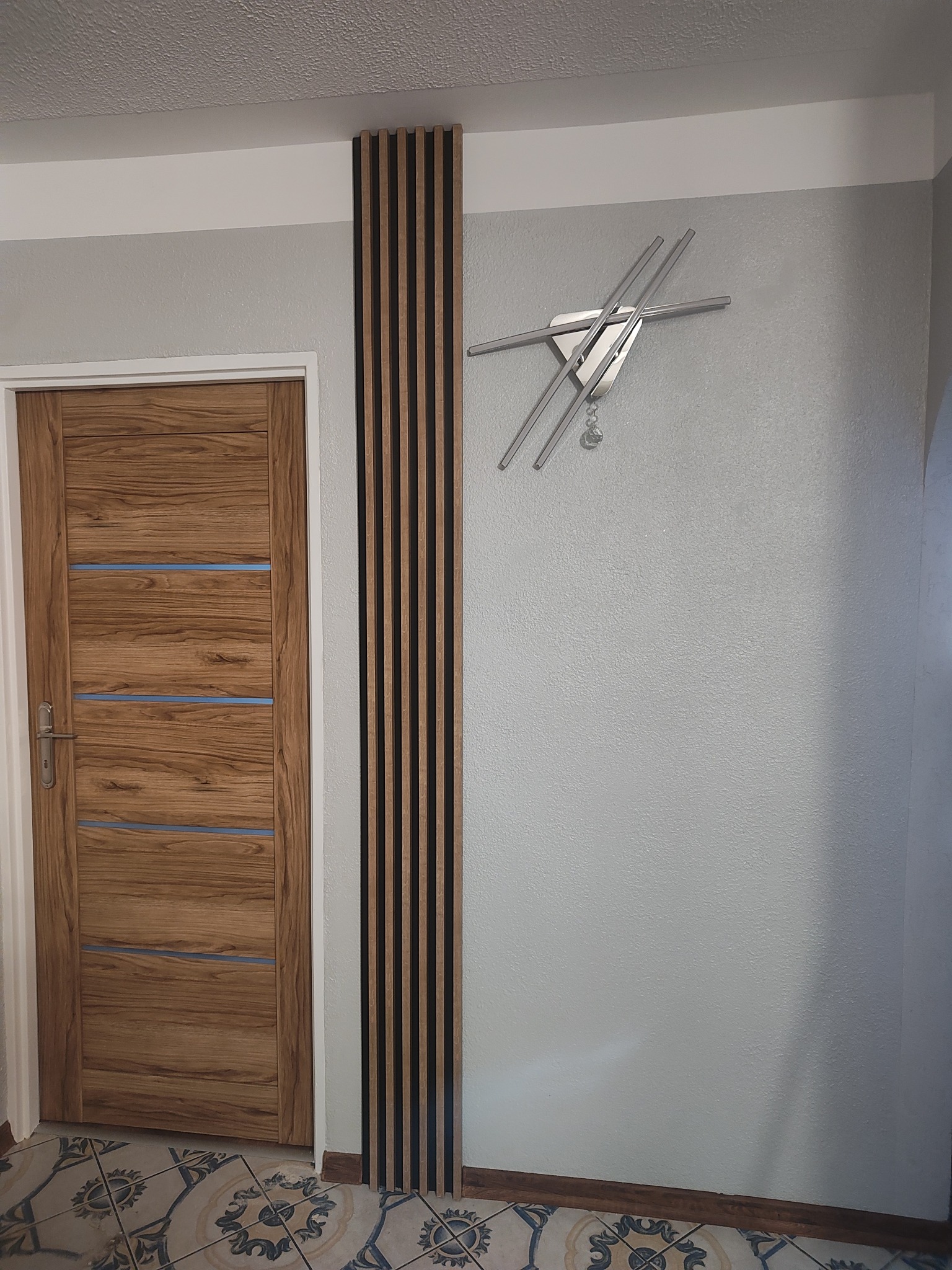 Nowoczesny przedpokój z drewnianymi lamelami na ścianie obok drzwi, uzupełniony o designerski zegar. Jasne ściany i podłoga z wzorem.