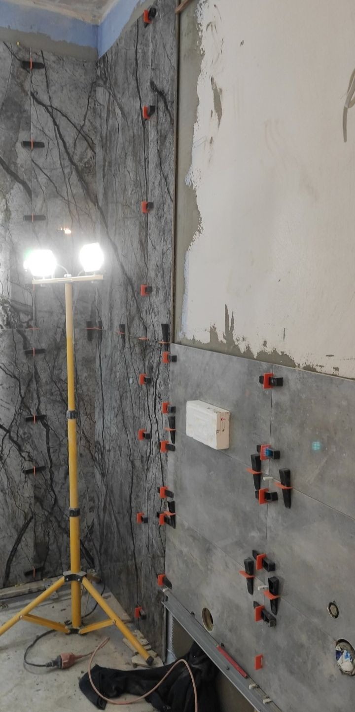 Układanie płytek imitujących marmur na ścianie, z użyciem poziomowania krzyżykami. Widoczne narzędzia i lampa oświetleniowa na trójnogu.