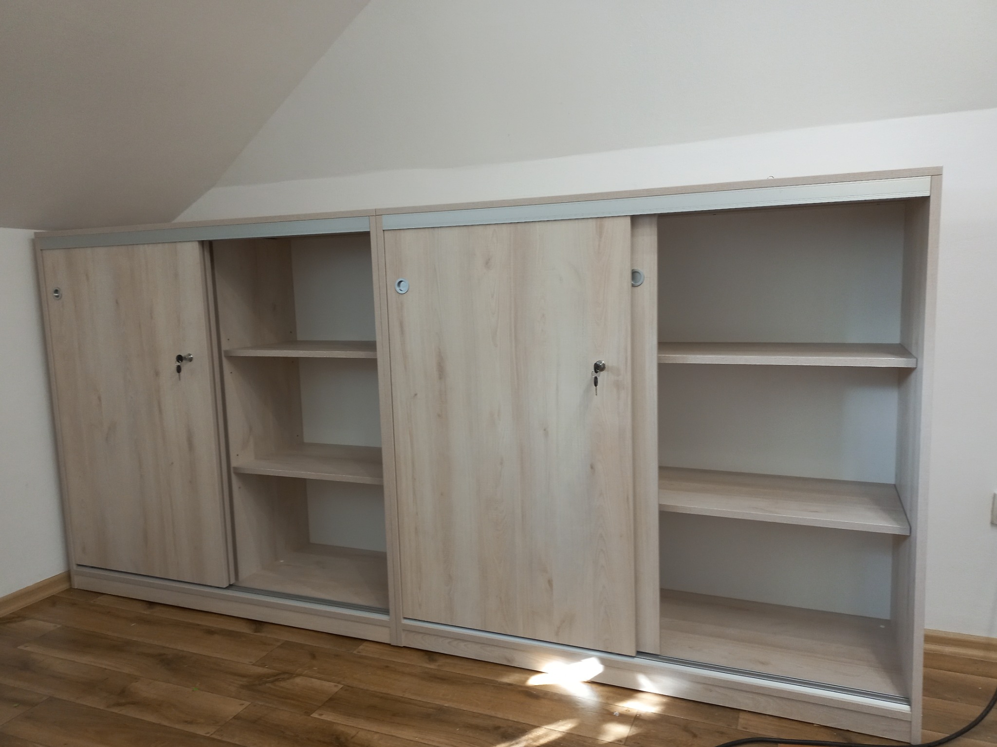 Jasna szafa wnękowa z przesuwnymi drzwiami, wykończona w odcieniach drewna, z zamkami w drzwiach i półkami w środku, na poddaszu z drewnianą podłogą.