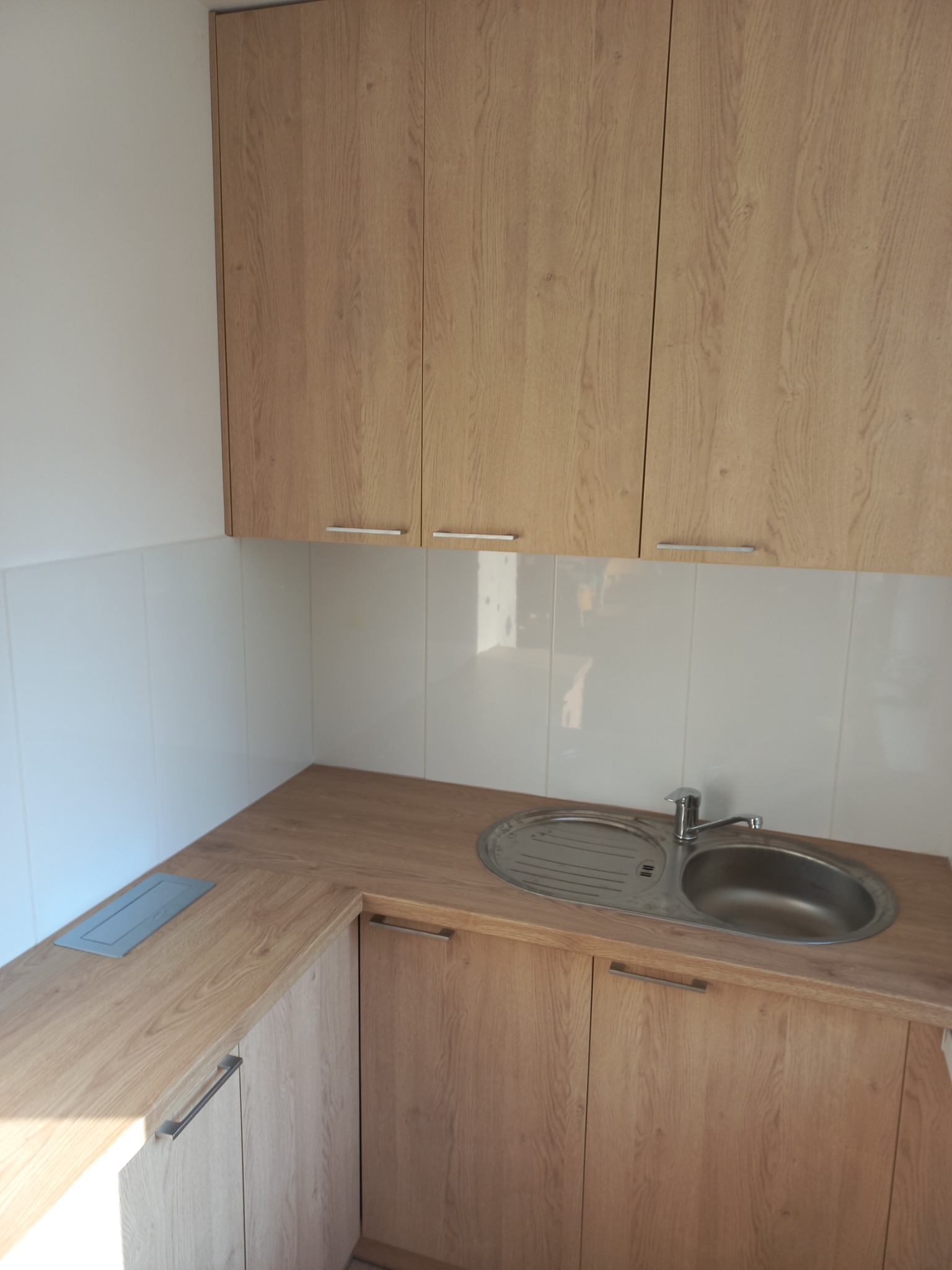 Jasna kuchnia z drewnopodobnymi frontami, widoczny zlew dwukomorowy i górne szafki z poziomymi uchwytami, białe płytki na ścianie.