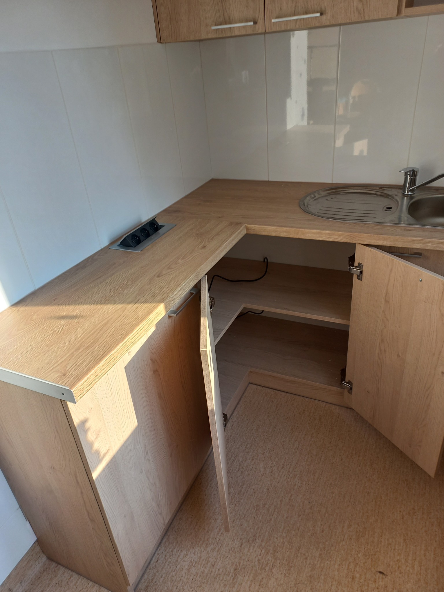 Jasna, drewnopodobna kuchnia na wymiar z otwartymi drzwiczkami szafki narożnej, widoczne gniazdka blatowe i dwukomorowy zlew ze stali nierdzewnej.