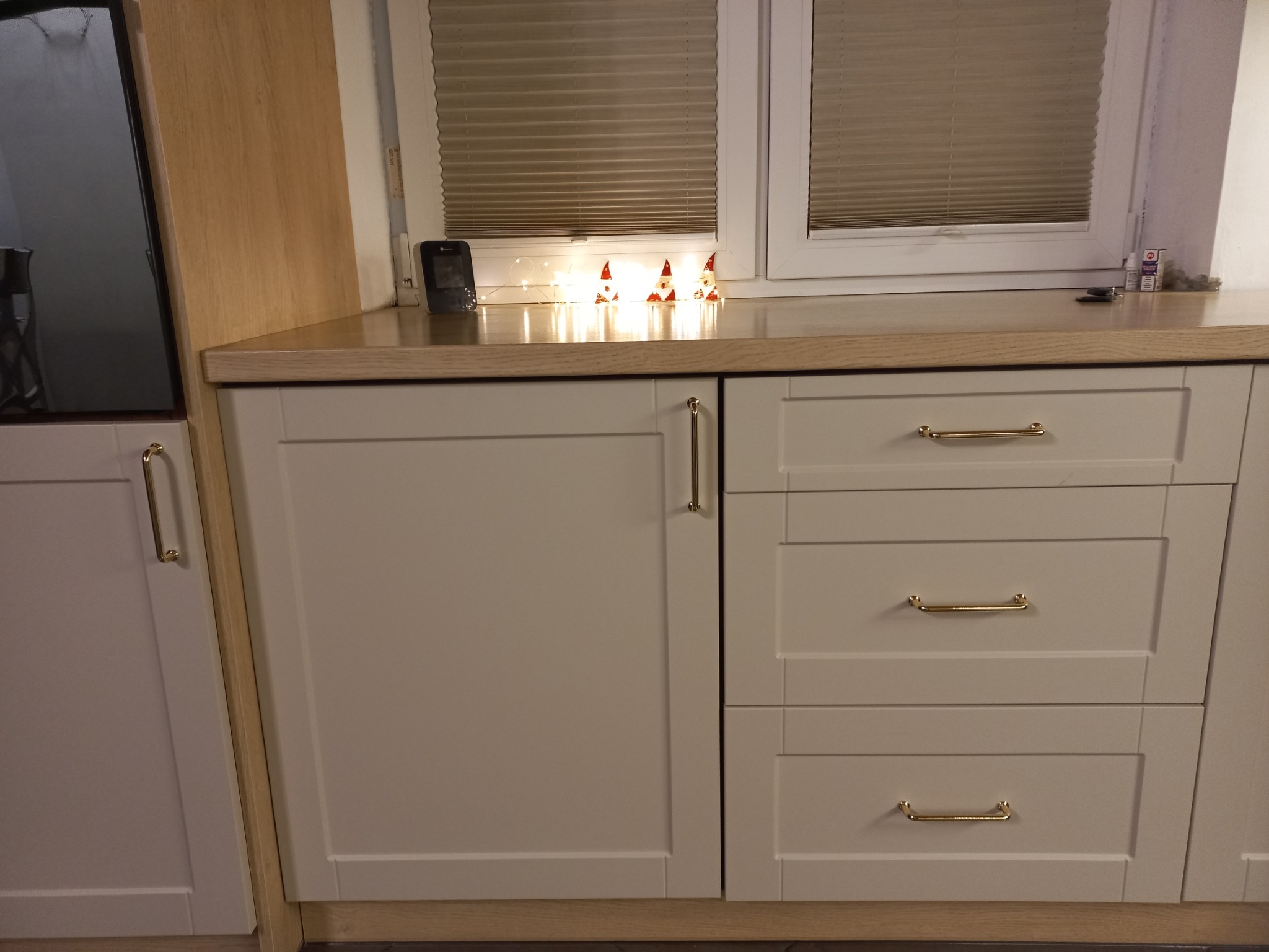 Fragment zabudowy kuchennej z białymi frontami, złotymi uchwytami i blatem w odcieniu jasnego drewna, ozdobiony świątecznymi figurkami gnomów i lampkami LED na tle okna z żaluzjami.