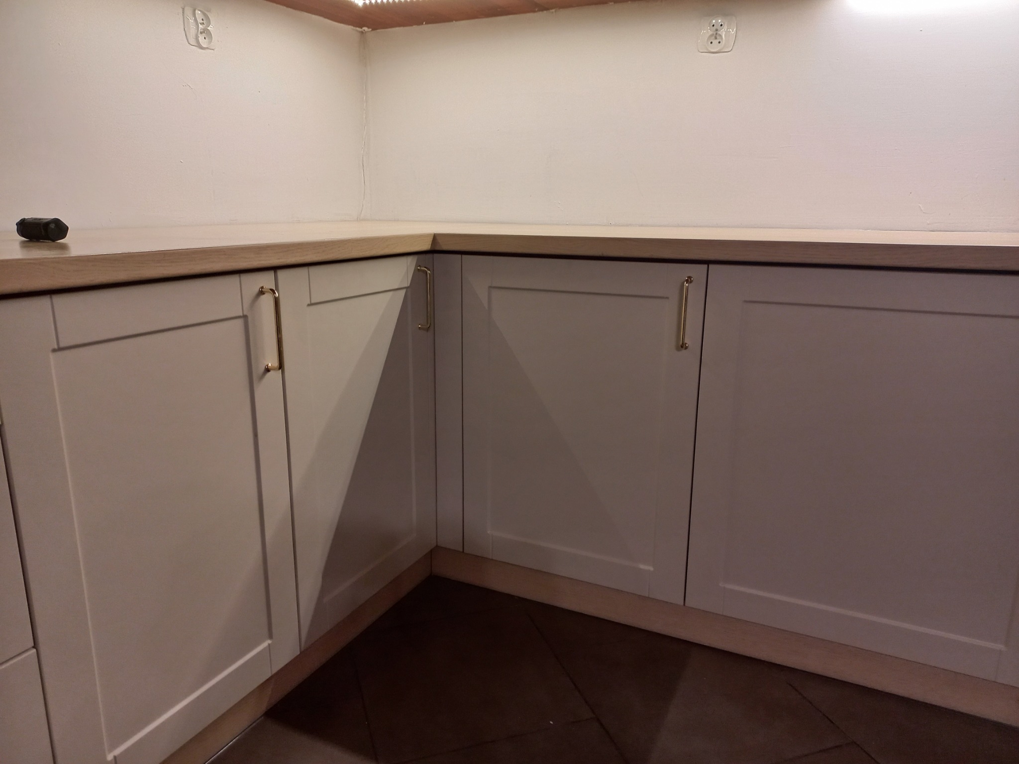 Fragment narożnej zabudowy kuchennej z szarymi frontami i drewnianym blatem, widoczny włącznik na blacie, gniazdka elektryczne na ścianie.