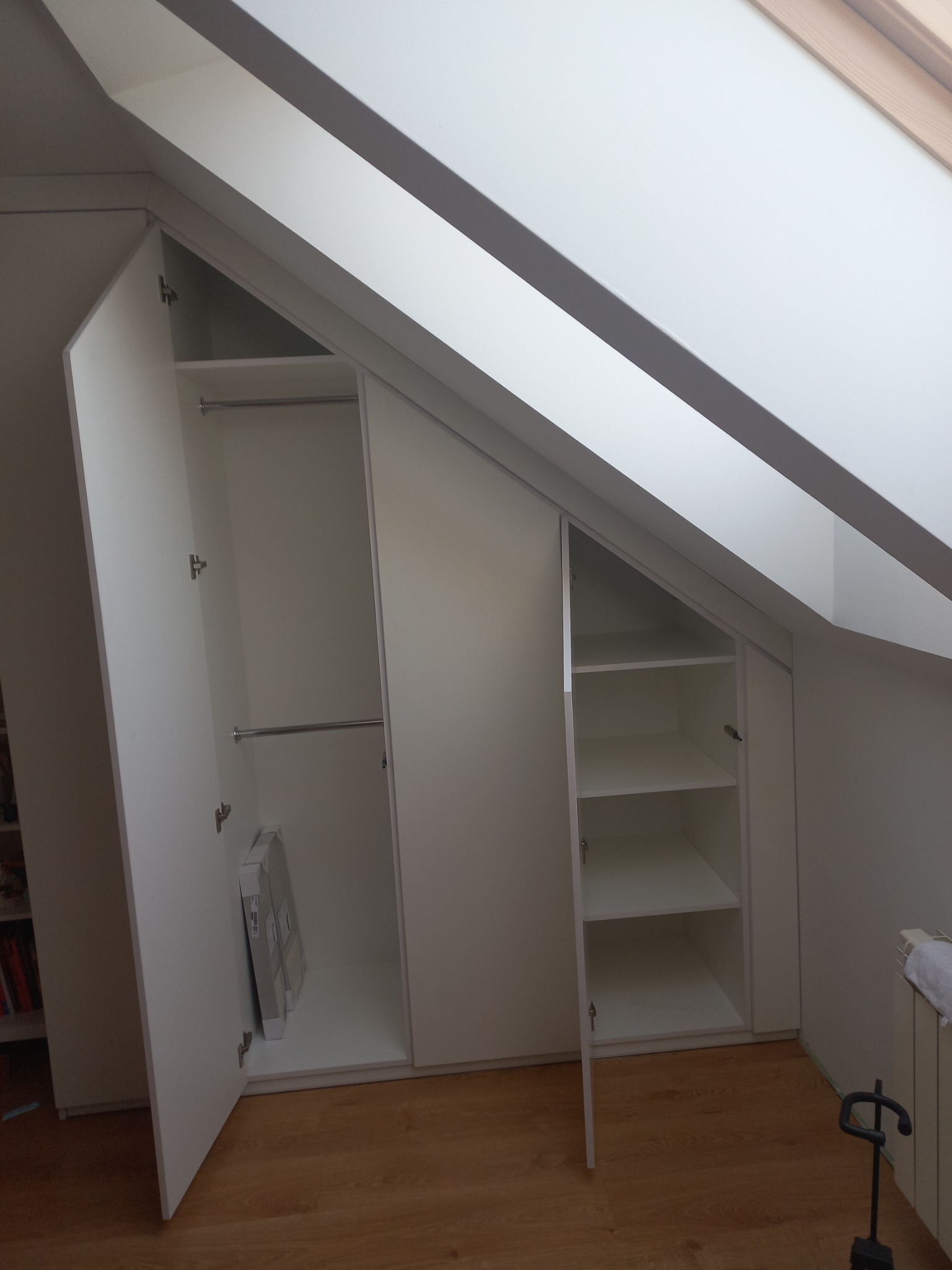 Biała szafa wnękowa z otwartymi drzwiami i półkami, dopasowana do skosu dachu, z widocznymi zawiasami i konstrukcją wewnętrzną.