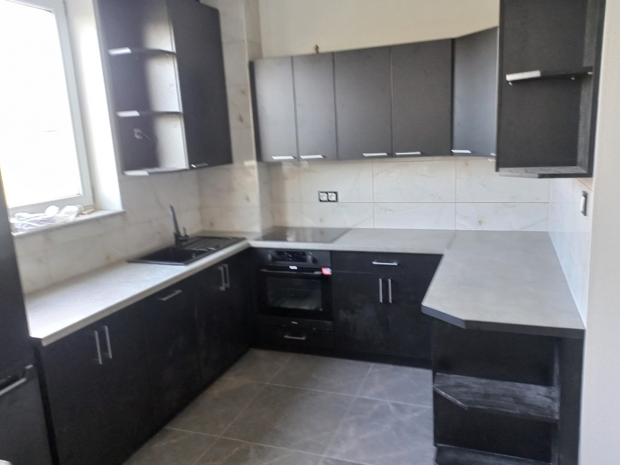 Nowoczesna, czarna kuchnia na wymiar z szarym blatem i płytkami imitującymi marmur. Widoczne górne szafki i sprzęt AGD. Ujęcie z perspektywy podłogi.