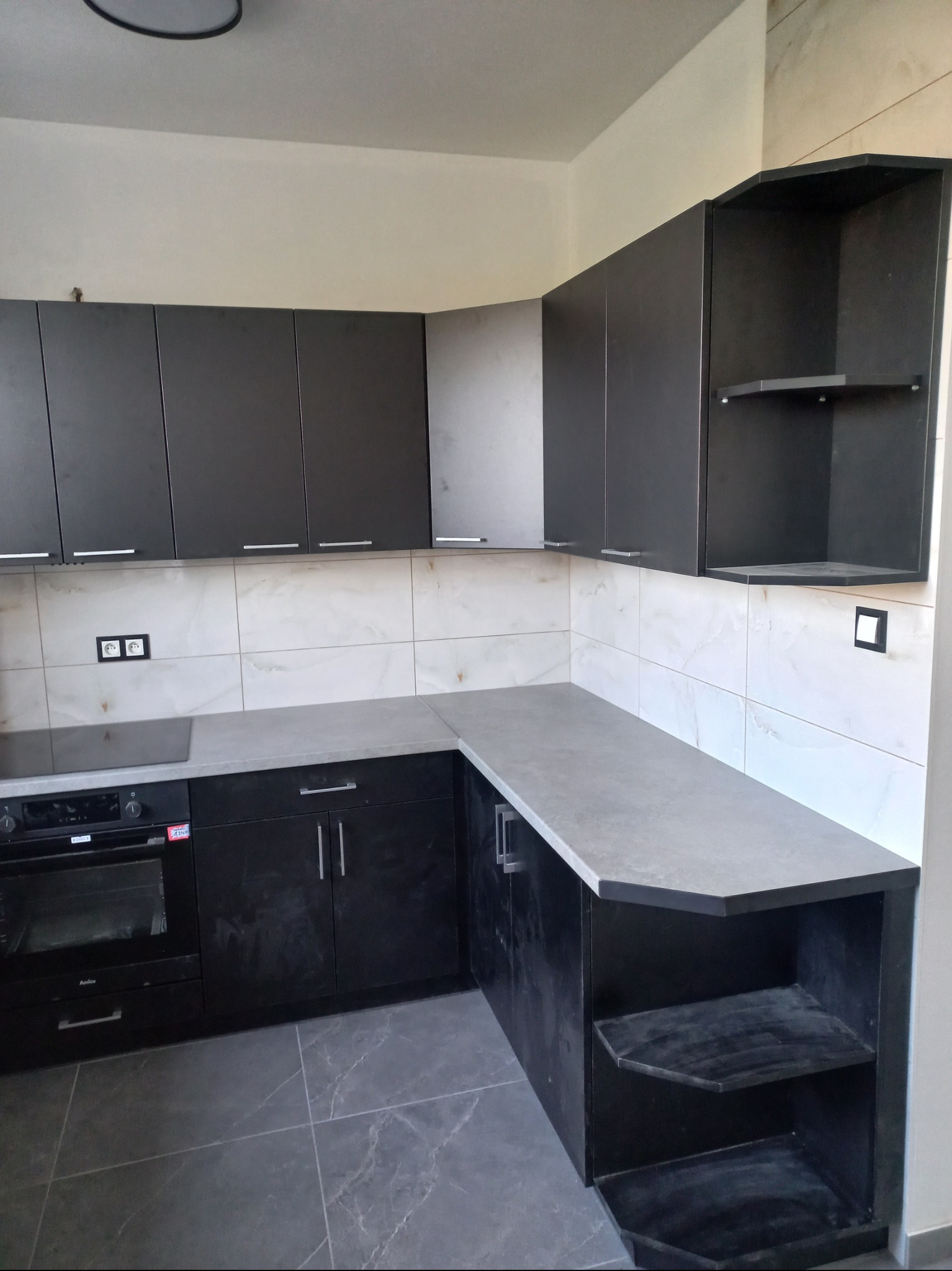 Nowoczesna, czarna kuchnia narożna z szarym blatem i płytkami imitującymi marmur. Widoczne gniazdka elektryczne i piekarnik. Dolna szafka z półkami.