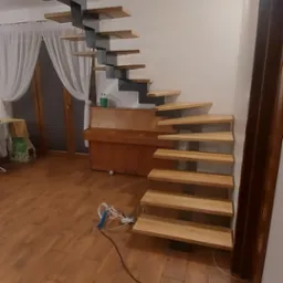 Nowoczesne schody z drewnianymi stopniami i metalową konstrukcją w jasnym wnętrzu, widoczne przewody narzędzi na podłodze.