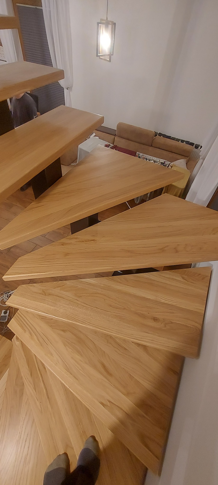 Spiralne schody z jasnego drewna, z czarnymi wspornikami, widziane z góry, z fragmentem osoby w tle i stopami w skarpetach w dolnym rogu.