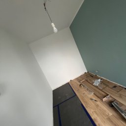 Usługi Remontowo Budowalne Bartłomiej Podliński - Wnętrze w trakcie prac: częściowo ułożone panele podłogowe, zabezpieczona folią część podłogi, ściany w odcieniach bieli i zieleni. Widoczne narzędzia i akcesoria.