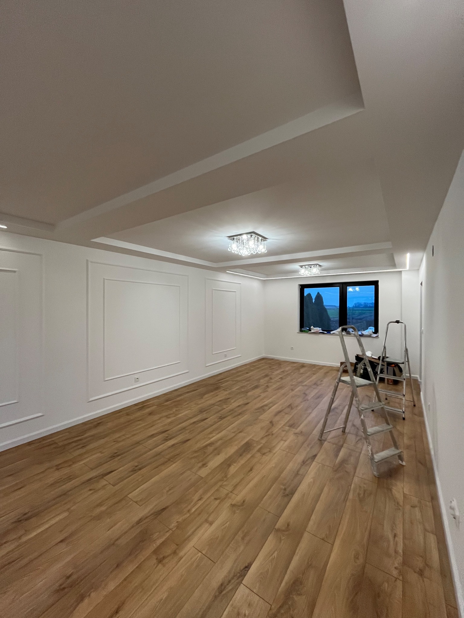 Jasne wnętrze po remoncie: drewnopodobna podłoga, białe ściany z dekoracyjnymi listwami, sufit z oświetleniem. Drabina malarska przy oknie.