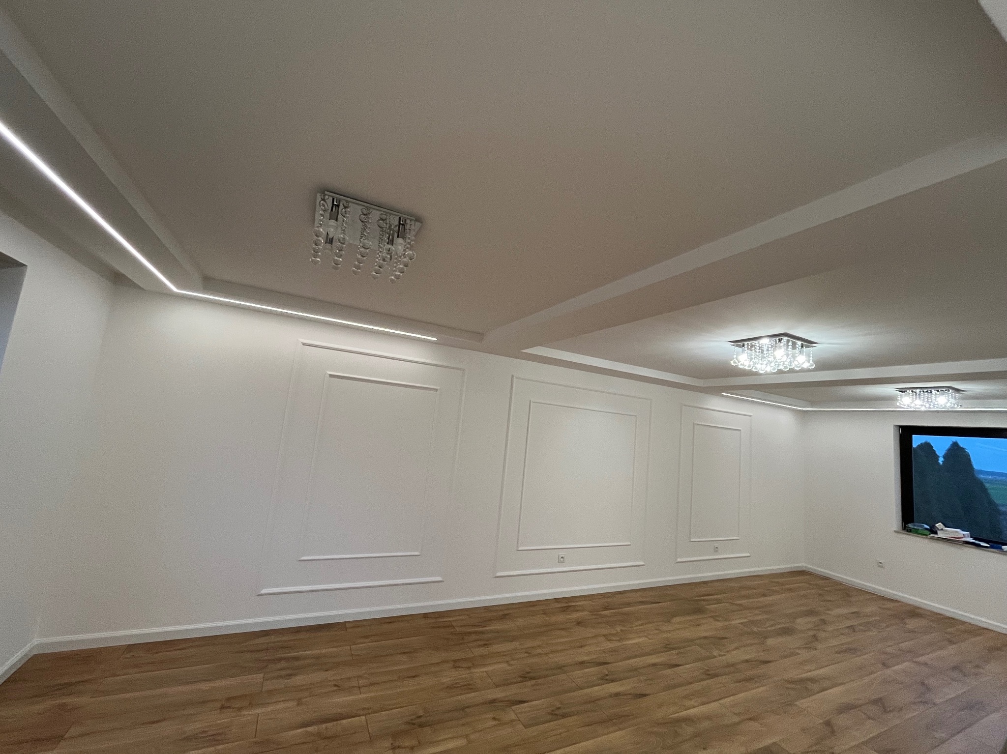 Jasne wnętrze po remoncie: drewniana podłoga, białe ściany z dekoracyjnymi listwami, sufit z oświetleniem LED i kryształowymi lampami. Widok na zewnątrz przez okno.