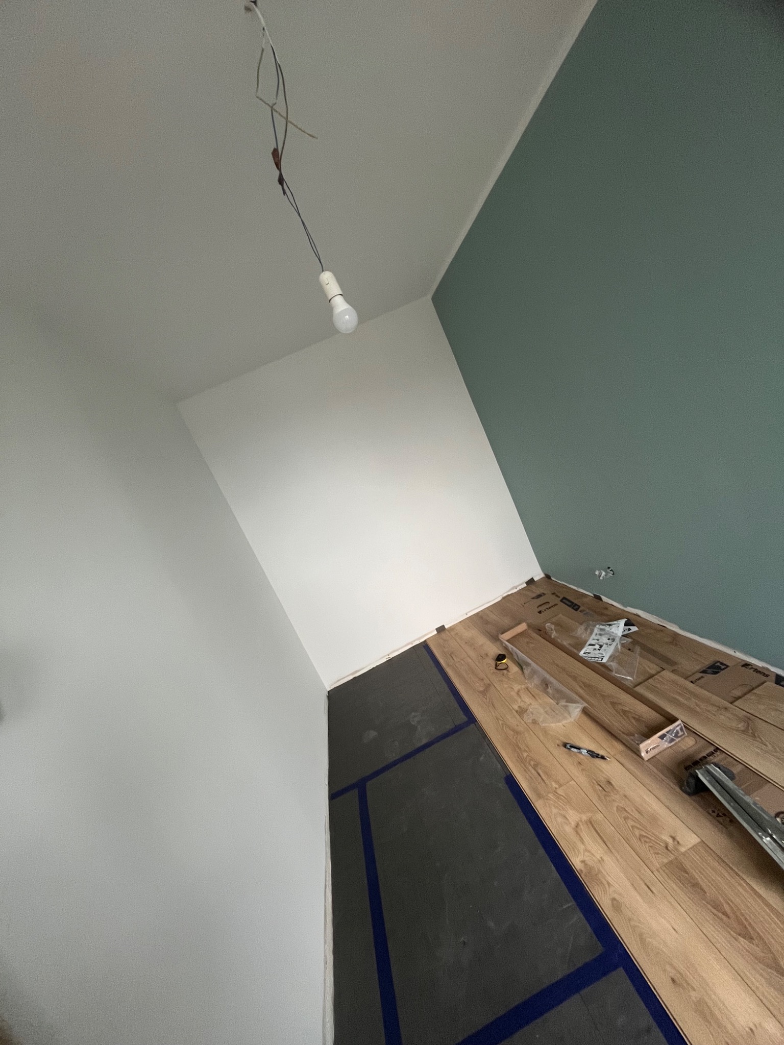 Wnętrze w trakcie prac: częściowo ułożone panele podłogowe, zabezpieczona folią część podłogi, ściany w odcieniach bieli i zieleni. Widoczne narzędzia i akcesoria.