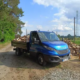 Niebieski samochód dostawczy Iveco z pełną skrzynią załadowaną pociętym drewnem na tle drzew i stosu drewna. Na pierwszym planie widoczna ziemista droga.