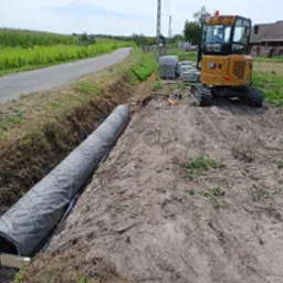 Żółta mini koparka CAT obok wykopu z rurą drenażową w szarej osłonie, kostka brukowa i droga asfaltowa w tle.