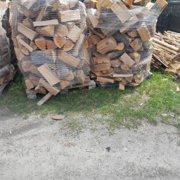 Krzysztof Kaczmarek - Dwa worki szczelnie owiniętego drewna kominkowego ułożone na paletach, obok sterta luźnych kawałków drewna, na piaszczystym podłożu z kępami trawy.