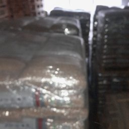 Krzysztof Kaczmarek - Stosy worków z pelletem, owinięte folią, składowane w magazynie przy oświetlonym oknie. Widoczne inne palety w tle.