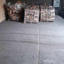 Krzysztof Kaczmarek - Trzy palety z ułożonym drewnem kominkowym, owinięte folią, w magazynie z betonową podłogą i ścianami z cegły.