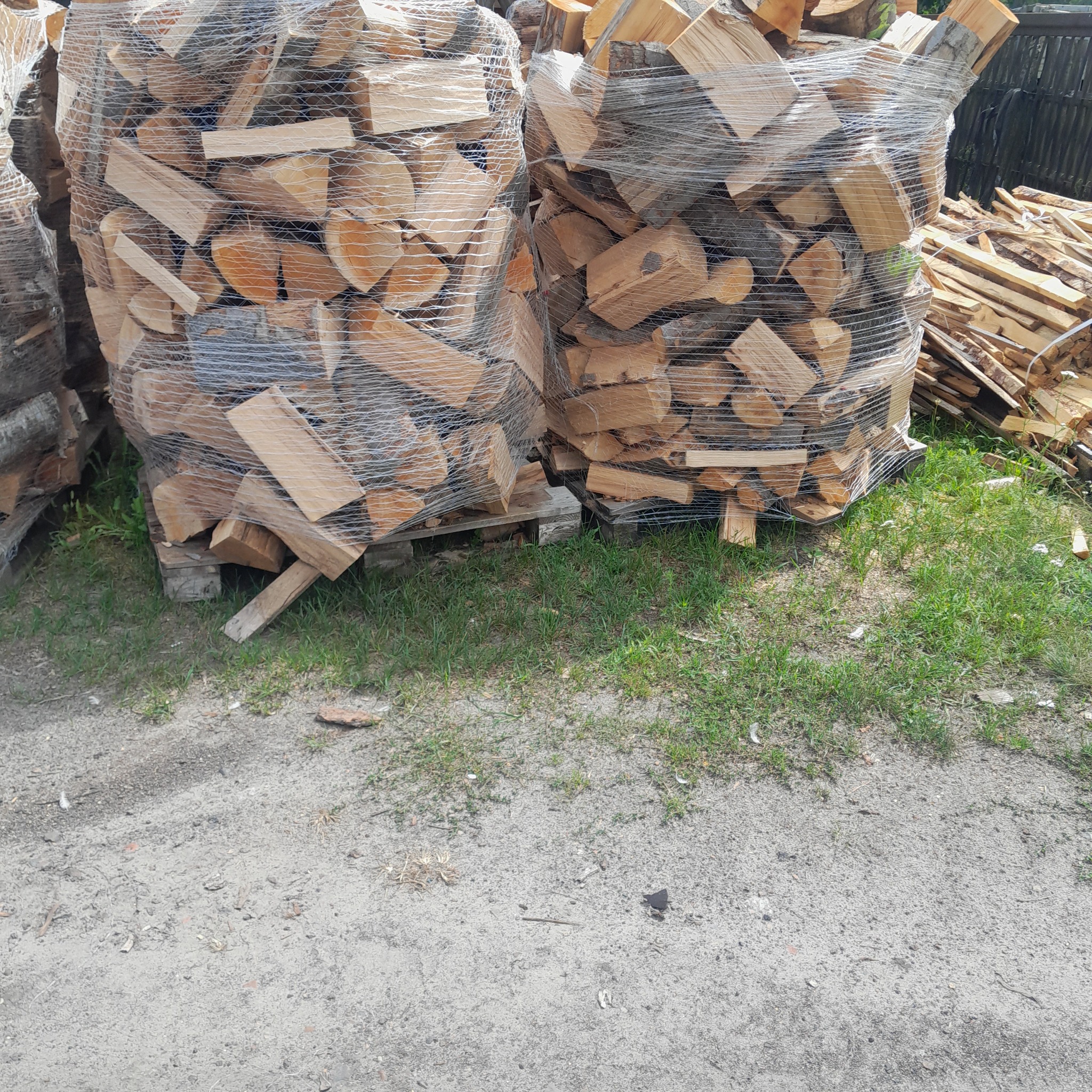 Dwa worki szczelnie owiniętego drewna kominkowego ułożone na paletach, obok sterta luźnych kawałków drewna, na piaszczystym podłożu z kępami trawy.