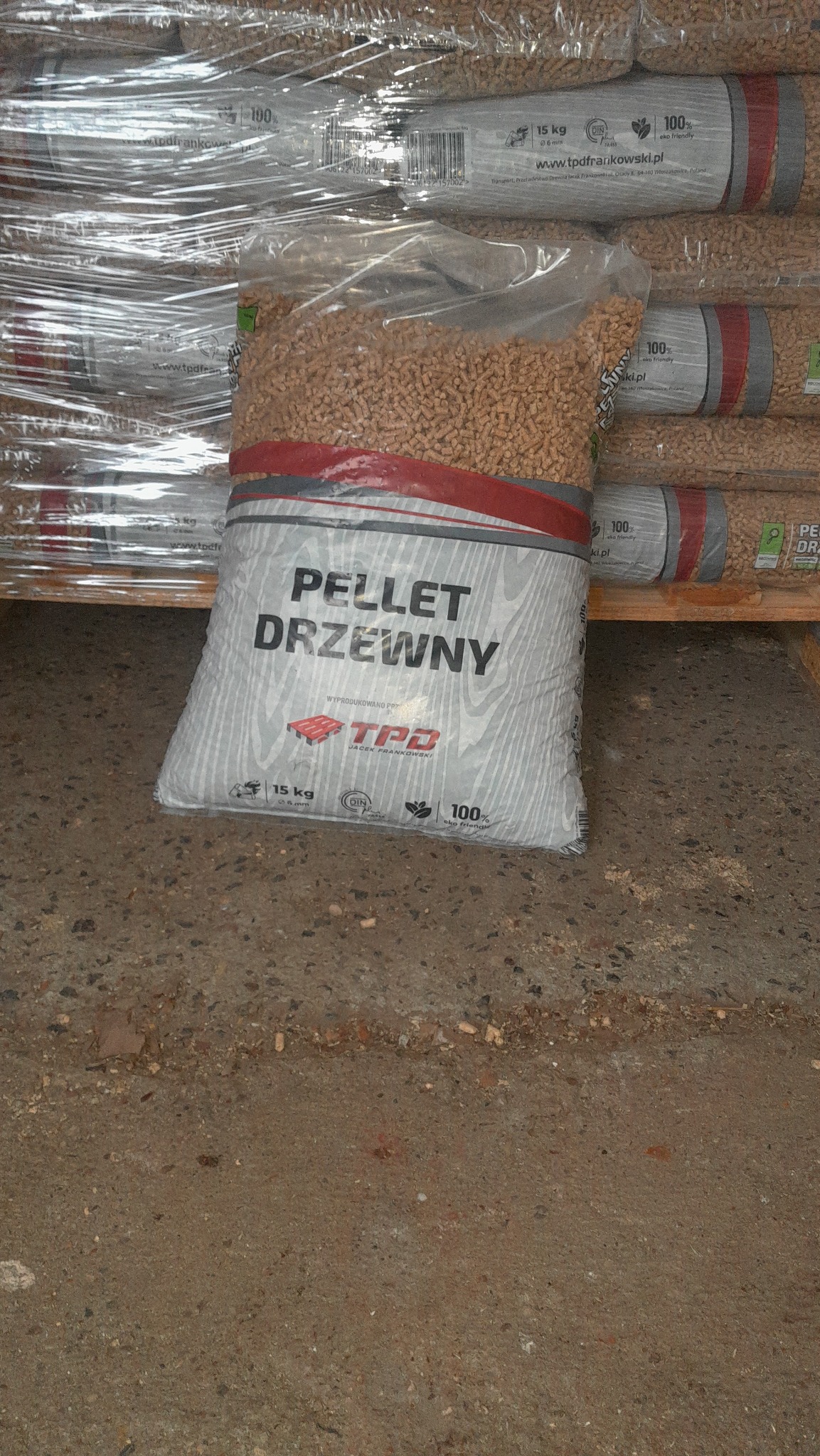 Wysoki stos worków z pelletem drzewnym, zapakowanych w przezroczystą folię, widocznych na palecie. Na pierwszym planie worek z napisem 'Pellet Drzewny' i logotypem producenta TPD.
