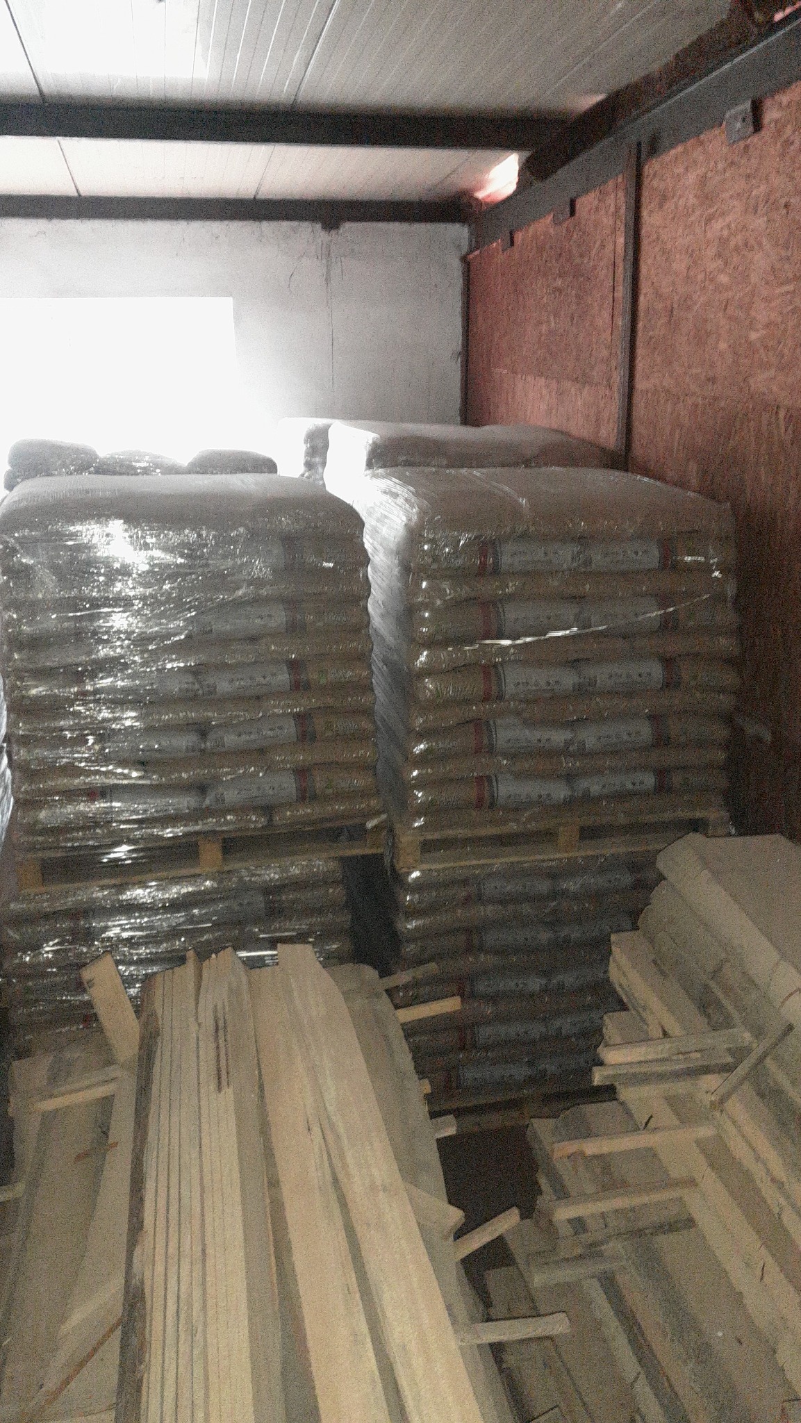 Stosy worków z pelletem na paletach, owinięte folią, składowane w magazynie obok stosu drewna.