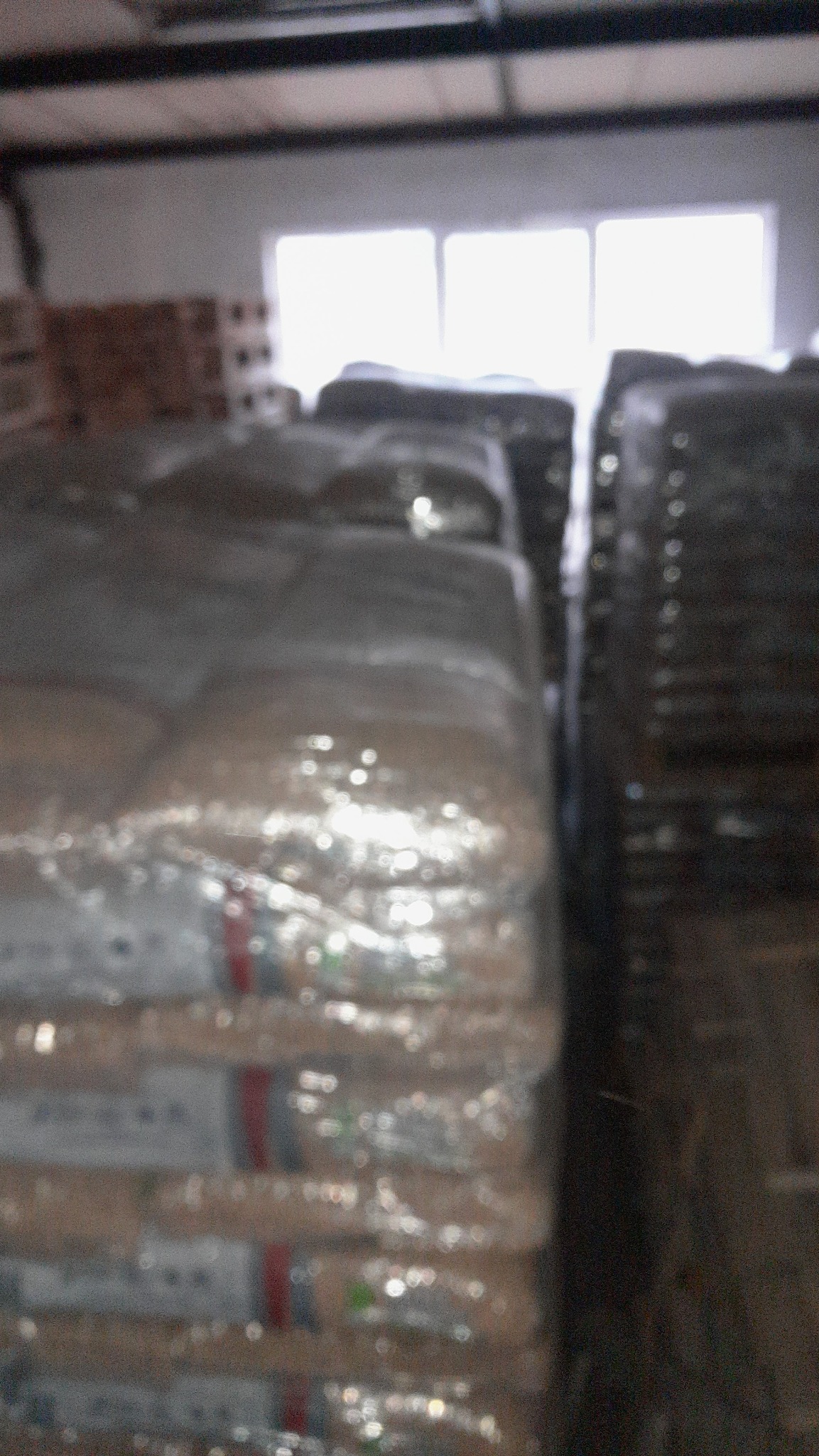 Stosy worków z pelletem, owinięte folią, składowane w magazynie przy oświetlonym oknie. Widoczne inne palety w tle.