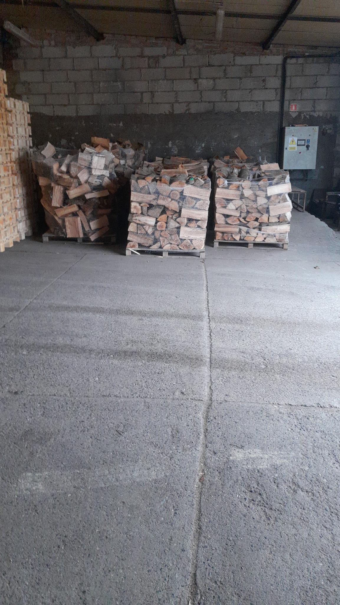 Trzy palety z ułożonym drewnem kominkowym, owinięte folią, w magazynie z betonową podłogą i ścianami z cegły.
