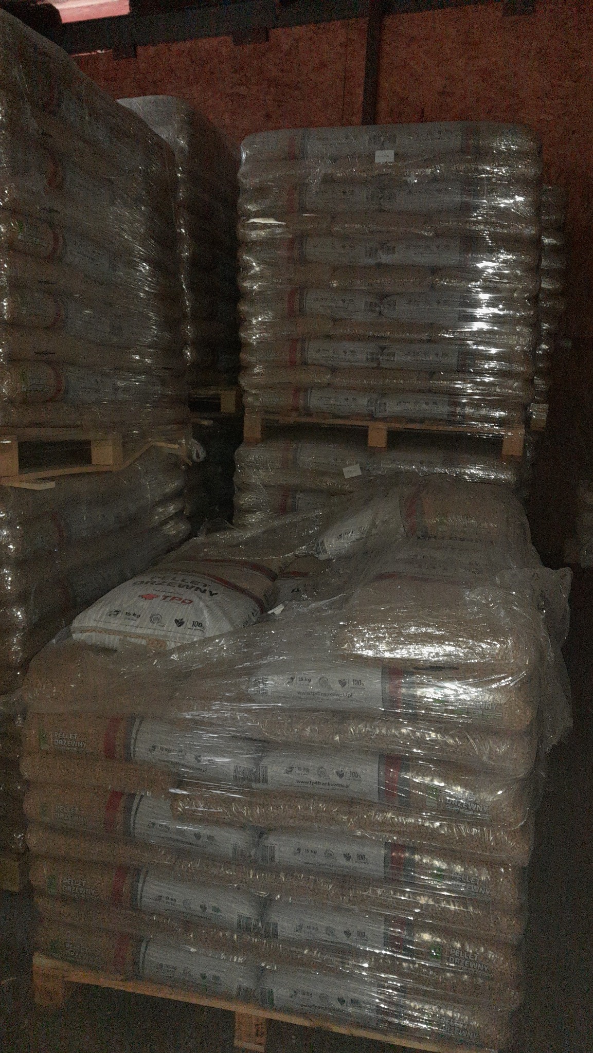 Stosy palet z workami pelletu drzewnego, owinięte folią, w magazynie. Widoczne etykiety na workach z informacjami o produkcie i wadze.