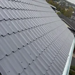 Szary dach z blachy trapezowej, widok z góry pod kątem, rynna wzdłuż krawędzi, zabudowa mieszkalna w tle pod pochmurnym niebem.