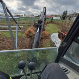 Widok z kabiny maszyny budowlanej podczas prac ziemnych na terenie z konstrukcjami szklarni i metalowymi ramami, operator w czarnych spodniach, widoczne dźwignie sterujące.