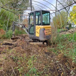 Żółto-szara minikoparka Volvo wewnątrz metalowej konstrukcji szklarni, częściowo zasłonięta przez krzewy i roślinność.