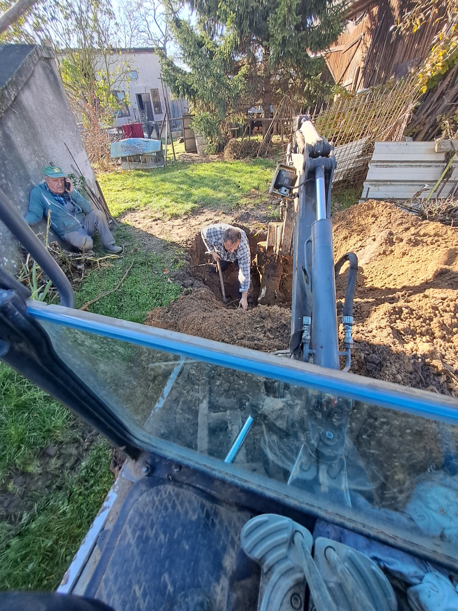 Widok z kabiny minikoparki na pracownika kopiącego dół łopatą, podczas gdy drugi pracownik rozmawia przez telefon siedząc obok wykopu.