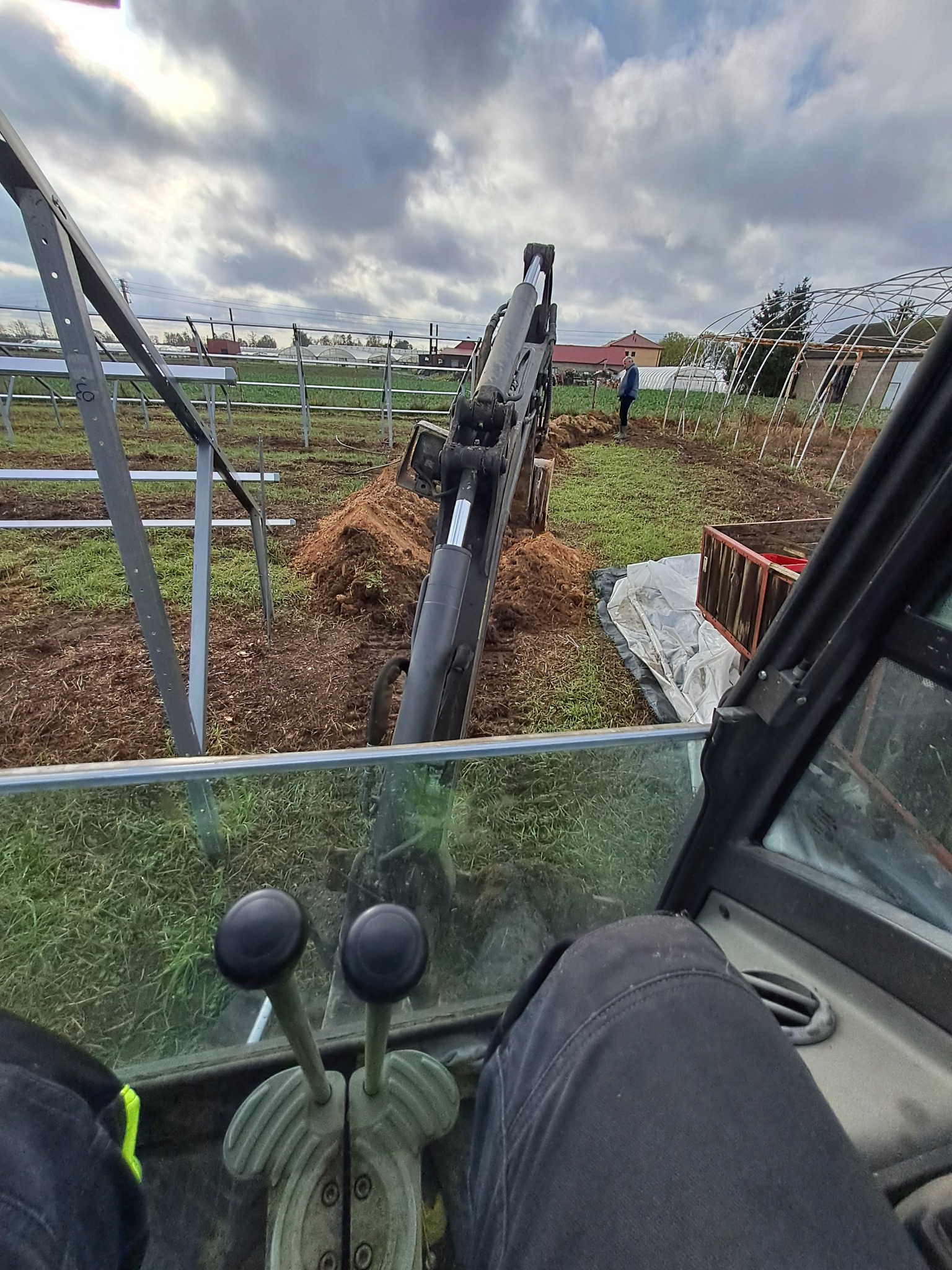Widok z kabiny maszyny budowlanej podczas prac ziemnych na terenie z konstrukcjami szklarni i metalowymi ramami, operator w czarnych spodniach, widoczne dźwignie sterujące.