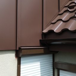 RAD-BUD Ekodomy Sp. z o. o. - Fragment budynku z brązową elewacją panelową i dachówką ceramiczną w odcieniach brązu, widoczne rynny i rolety zewnętrzne.