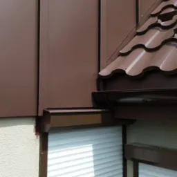 Fragment budynku z brązową elewacją panelową i dachówką ceramiczną w odcieniach brązu, widoczne rynny i rolety zewnętrzne.